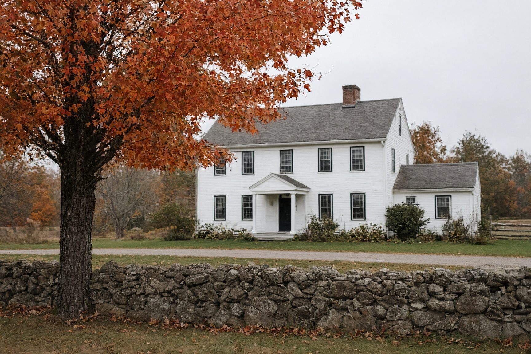 White colonial house behind a fieldstone wall, with an orange-leafed maple in the yard