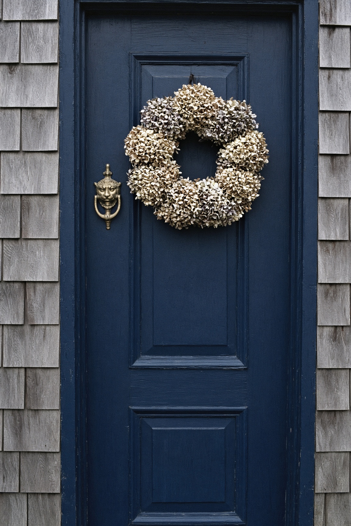 Navy front door with a brass lion-head knocker and a dried hydrangea wreath, set in weathered gray shingles